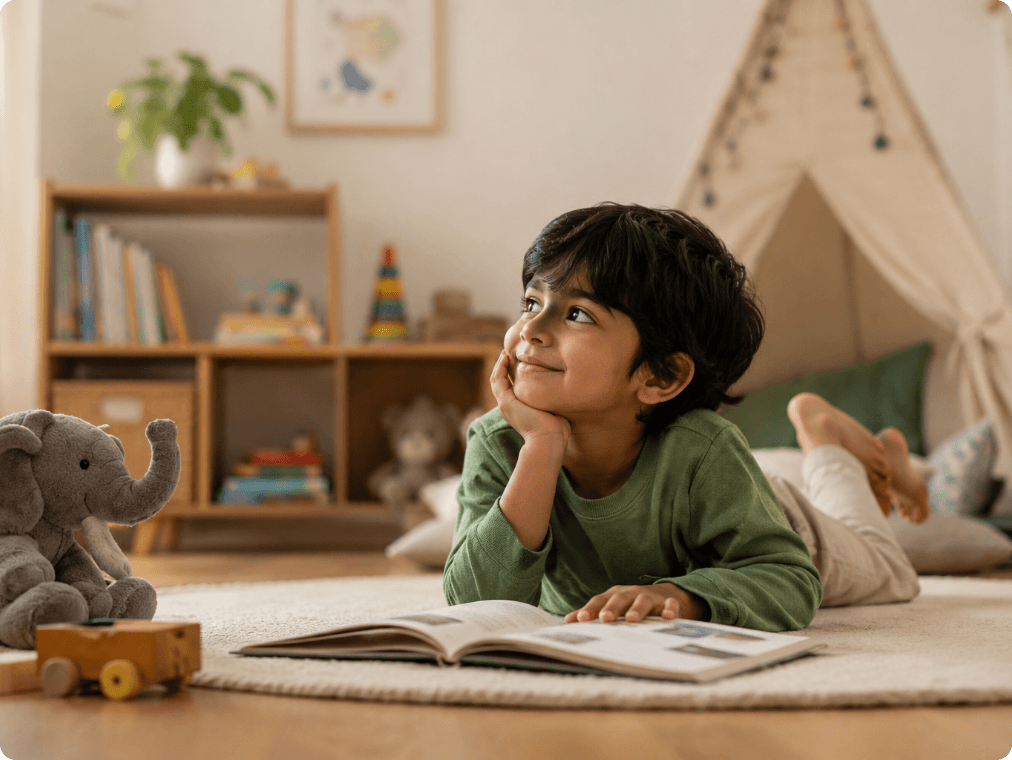 A child lying on a rug, resting their chin on their hand and looking up with a smile beside an open book